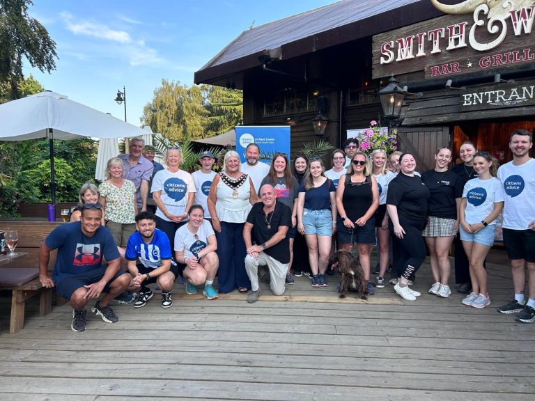 group of people smiling outside of a pub after completing a 10km walk, mix of genders, some kneeling down and others standing up behind them. some with white t shirts that say Citizens Advice. A dog also in the photo at the front, the dog is brown and fluffy
