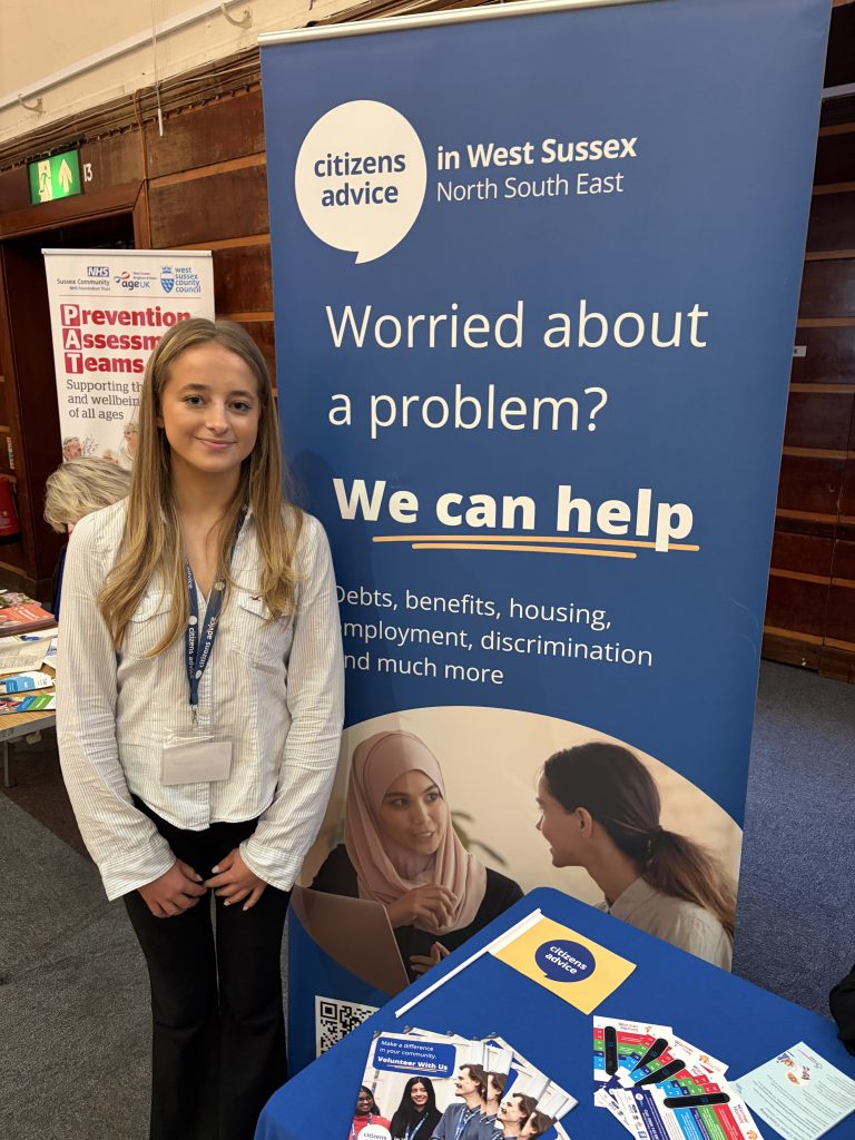 A young girl, blondish hair, light blue striped shirt, black trousers, standing in from of a Citizens Advice West Sussex banner saying We can help. Her hands are just in front of her an there is a table beside her with leaflets about Citizens Advice West Sussex